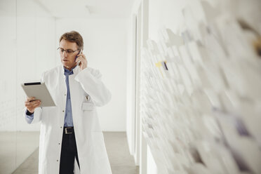 Mature man in lab coat on phone looking at digital tablet - MFF001521