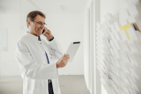 Lächelnder reifer Mann im Laborkittel am Telefon mit Blick auf ein digitales Tablet, lizenzfreies Stockfoto