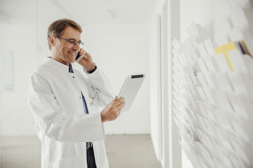 Lächelnder reifer Mann im Laborkittel am Telefon mit Blick auf ein digitales Tablet - MFF001523