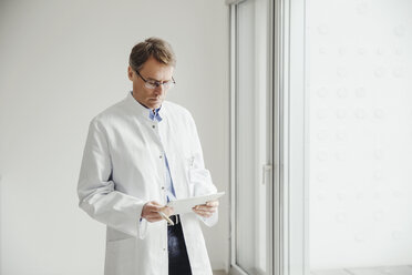 Mature man in lab coat using digital tablet at the window - MFF001536