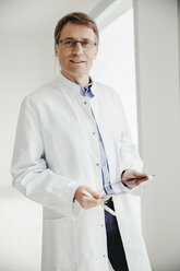 Smiling mature man in lab coat holding digital tablet at the window - MFF001537