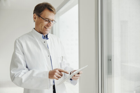 Lächelnder reifer Mann im Laborkittel, der ein digitales Tablet am Fenster benutzt - MFF001538