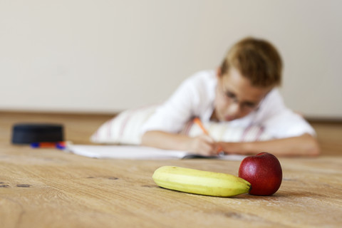 Junge macht Hausaufgaben auf Holzboden mit Banane und Apfel im Vordergrund, lizenzfreies Stockfoto