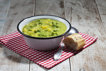 Enamel bowl of pea stew, spoon and sliced baguette on cloth and wood - CSF024977