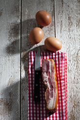 Speck und Küchenmesser auf Handtuch und drei braune Hühnereier auf Holz - CSF025011