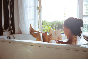 Woman relaxing in bathtub reading book - MBEF001335