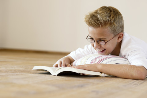 Lächelnder Junge, der auf dem Holzboden liegt und ein Buch liest, lizenzfreies Stockfoto