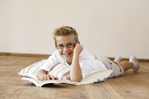 Porträt eines lächelnden Jungen, der mit einem Buch auf dem Holzboden liegt, lizenzfreies Stockfoto