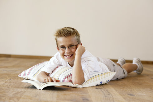 Porträt eines lächelnden Jungen, der mit einem Buch auf dem Holzboden liegt - LBF001098