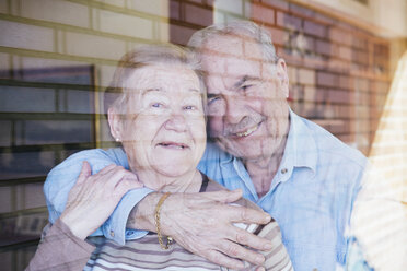 Elderly couple embracing at home looking through window - GEMF000136