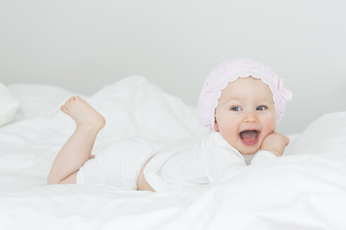 Laughing baby girl with cap lying on bed - JTLF000099