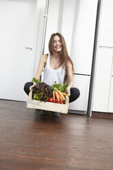 Glückliche junge Frau in der Küche mit einer Kiste voll frischem Gemüse - RHF000691