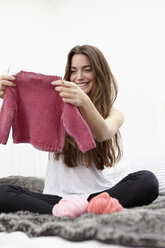 Young woman sitting on bed knitting kid's jumper - RHF000698