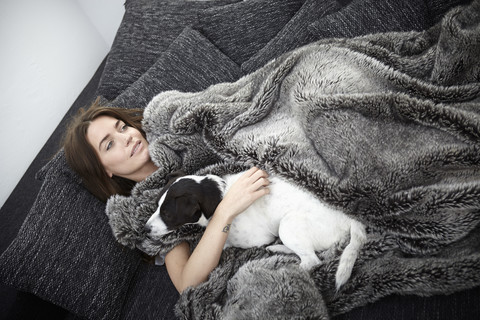 Junge Frau, eingewickelt in eine Pelzdecke, entspannt auf der Couch mit einem Hund, lizenzfreies Stockfoto