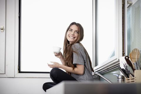 Junge Frau in der Küche, die am Fenster sitzt und Kaffee trinkt, lizenzfreies Stockfoto