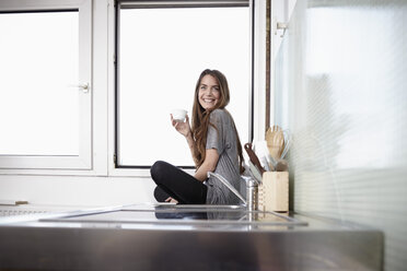 Junge Frau in der Küche, die am Fenster sitzt und Kaffee trinkt - RHF000737