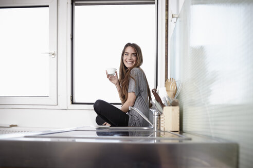 Young woman in kitchen sitting at window drinking coffee - RHF000737