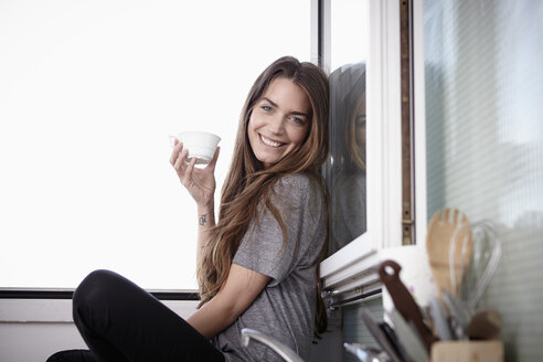 Young woman in kitchen sitting at window drinking coffee - RHF000738