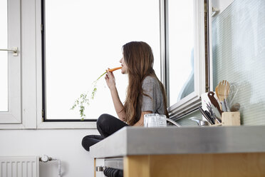 Junge Frau in der Küche, die am Fenster sitzt und Karotten isst - RHF000739