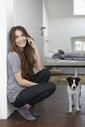 Junge Frau mit Hund zu Hause beim Telefonieren - RHF000742