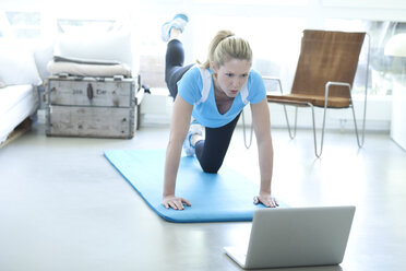 Woman looking at laptop exercising on gym mat in living room - MAEF010078