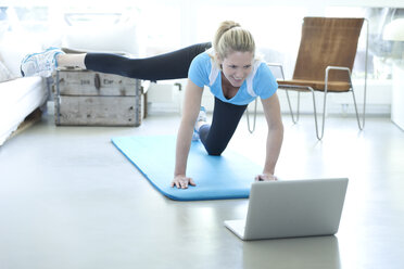 Woman looking at laptop exercising on gym mat in living room - MAEF010079