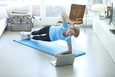 Woman looking at laptop exercising on gym mat in living room - MAEF010082