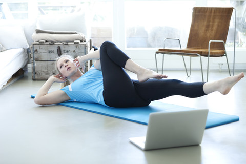 Frau mit Laptop, die auf einer Gymnastikmatte im Wohnzimmer trainiert, lizenzfreies Stockfoto