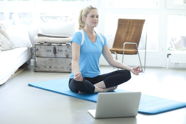 Frau mit Laptop übt Yoga auf Gymnastikmatte im Wohnzimmer - MAEF010084