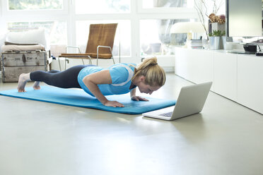Woman with laptop exercising on gym mat in living room - MAEF010089