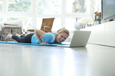 Woman with laptop exercising on gym mat in living room - MAEF010090