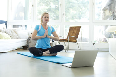 Frau mit Laptop übt Yoga auf Gymnastikmatte im Wohnzimmer - MAEF010092