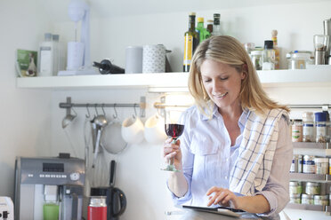 Smiling woman in kitchen drinking glass of red wine and using digital tablet - MAEF010112