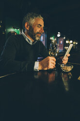Smiling man sitting at counter of a pub using smartphone - MBEF001356