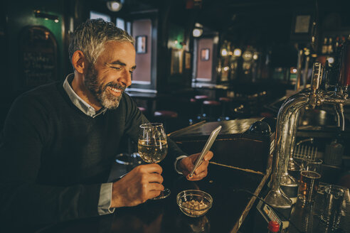 Portrait of smiling man sitting at counter of a pub using smartphone - MBEF001357