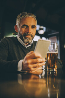Mann sitzt am Tresen einer Kneipe und schaut auf sein Smartphone - MBEF001363