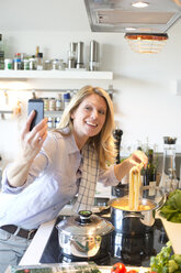 Smiling woman in kitchen cooking spaghetti taking a selfie - MAEF010162