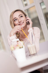 Lächelnde blonde Frau sitzt in einem Café mit einem Glas Latte Macchiato und telefoniert - JUNF000278