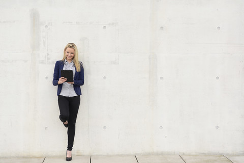 Lächelnde Geschäftsfrau mit digitalem Tablet, die sich an eine Betonwand lehnt, lizenzfreies Stockfoto