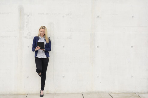 Lächelnde Geschäftsfrau mit digitalem Tablet, die sich an eine Betonwand lehnt - BFRF001035