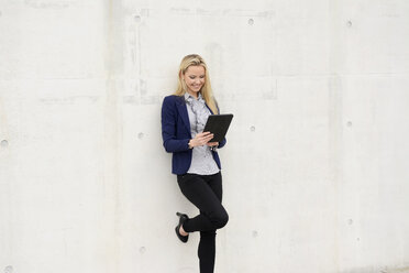 Lächelnde Geschäftsfrau mit digitalem Tablet, die sich an eine Betonwand lehnt - BFRF001036