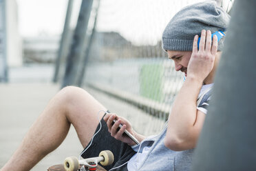 Young man with skateboard listening music from smartphone - UUF003856