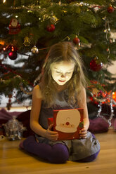 Little girl sitting in front of Christmas tree watching Christmas present - SARF001615