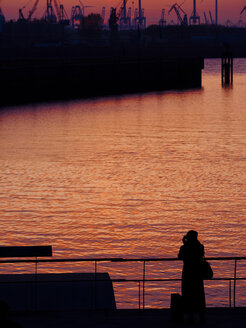 Deutschland, Hamburg, Hamburger Hafen, junge Fotografin steht am Elbufer bei Sonnenuntergang - KRPF001412