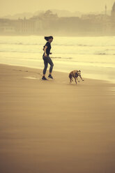 Spanien, Gijon, Frau joggt am Strand, schaut auf laufenden Hund - MGOF000190