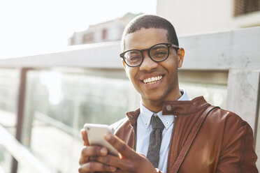 Porträt eines lächelnden Geschäftsmannes mit Smartphone, der eine Lederjacke und eine Brille trägt - EBSF000518