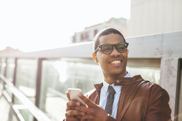 Portrait of smiling businessman with smartphone wearing leather jacket and glasses - EBSF000519