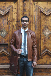 Portrait of businessman wearing leather jacket and glasses in front of wooden door - EBSF000528