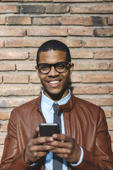 Portrait of smiling businessman with smartphone wearing leather jacket and glasses - EBSF000532