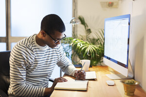 Young creative man sitting at desk writing in his notebook - EBSF000541